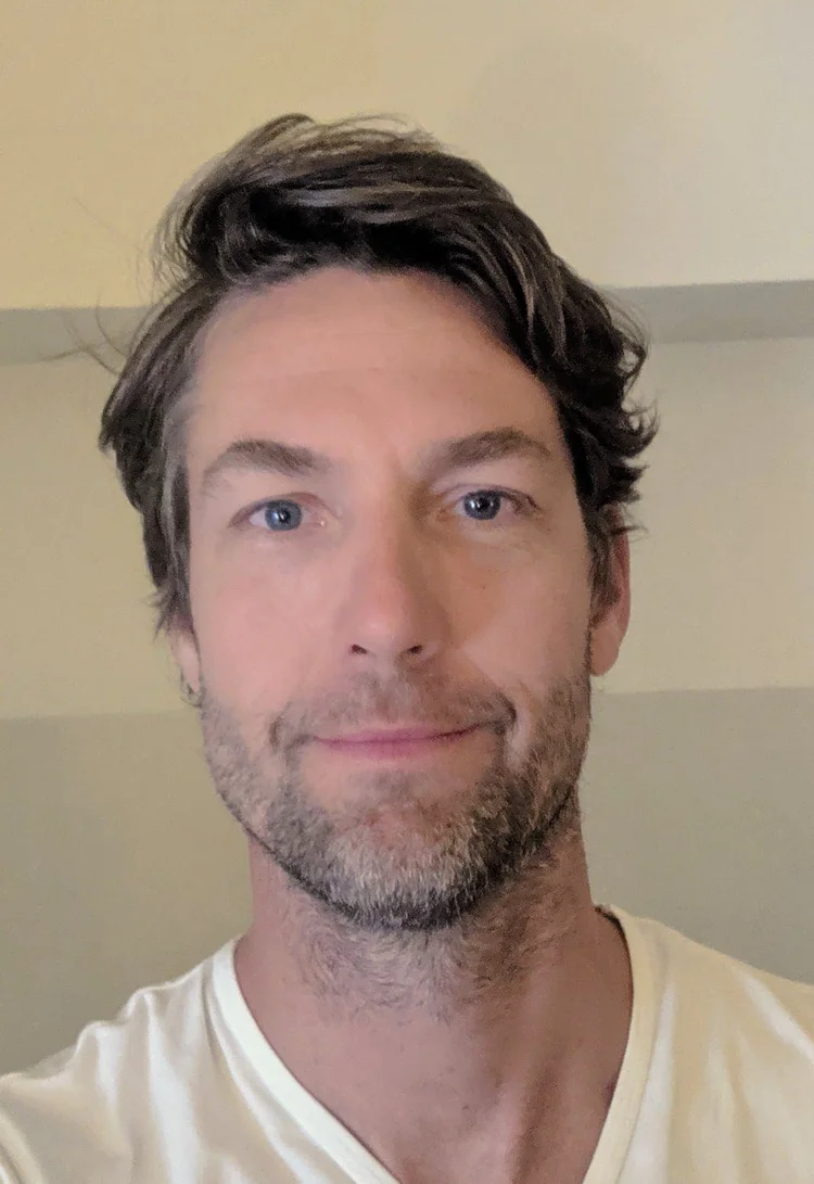 Un homme à la peau claire, aux cheveux bruns courts et ondulés, aux yeux clairs, vêtu d'une chemise blanche, sourit légèrement à la caméra.