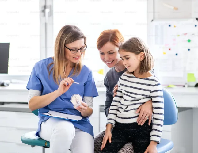 Dentiste ou hygiéniste dentaire donnant des instructions de brossage à un jeune patient, sous le regard de sa mère, dans un cabinet dentaire.